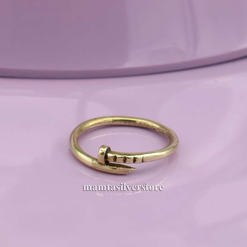 May include: A gold-toned ring with a nail design. The nail is bent at a 90-degree angle and has a textured surface.