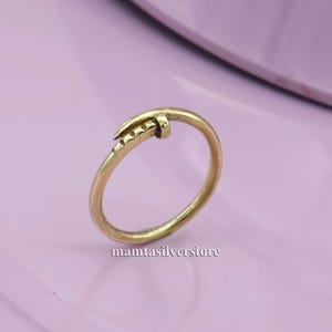 May include: A gold-toned ring with a nail design. The nail is bent into a curve and has a textured head.
