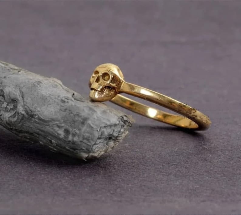May include: A gold skull ring featuring a detailed skull design. The ring is crafted from smooth, shiny metal and has a simple band.