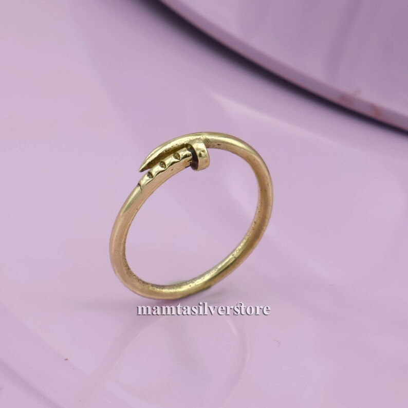 May include: A gold-colored nail ring with a simple design. The nail is bent into a ring shape and has a textured surface.