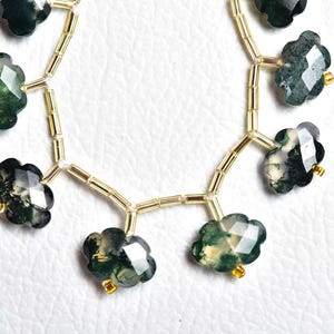May include: A strand of faceted, cloud-shaped moss agate beads in shades of dark green and translucent white, interspersed with small, cylindrical gold-toned spacer beads. The beads are arranged in a curved line on a white textured surface.
