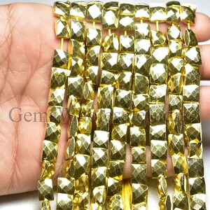 May include: Strands of golden, faceted square beads. The beads have a metallic sheen and are strung together. The image shows multiple strands, with the beads held in a hand. The beads are approximately 0.5 inches square.