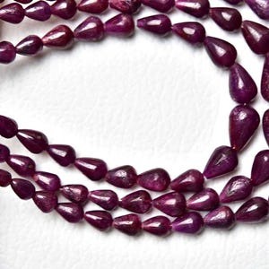 Op de afbeelding: Een streng diep magenta, traanvormige edelsteenkralen gerangschikt in een meerlaagse lus. De kralen hebben een gladde, gepolijste afwerking en zijn geregen aan een dun, lichtgekleurd koord, met een consistente donkere paars-rode kleur.