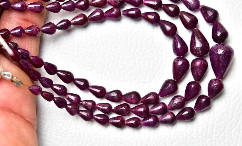 Op de afbeelding: Een streng diep magenta, traanvormige edelsteenkralen gerangschikt in een meerlaagse lus. De kralen hebben een gladde, gepolijste afwerking en zijn geregen aan een dun, lichtgekleurd koord, met een consistente donkere paars-rode kleur.