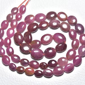 May include: Strand of oval-shaped, polished ruby gemstones in varying shades of red and pink. The gemstones are strung together with a red string, creating a necklace or bracelet component. The beads have a glossy finish.