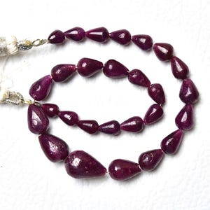 May include: A strand of deep magenta, pear-shaped ruby beads arranged in a circular pattern. The beads are polished and vary slightly in size, featuring two off-white, silky tassels attached to the ends of the string with silver-toned metal caps.