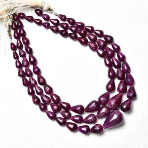 Op de afbeelding: Een ketting met drie strengen bestaande uit diep magenta, peervormige edelsteenkralen. De kralen zijn gefacetteerd en in een trapsgewijs patroon gerangschikt, bovenaan vastgezet met een lichtgekleurd kwastje en metalen onderdelen voor een gelaagd effect.