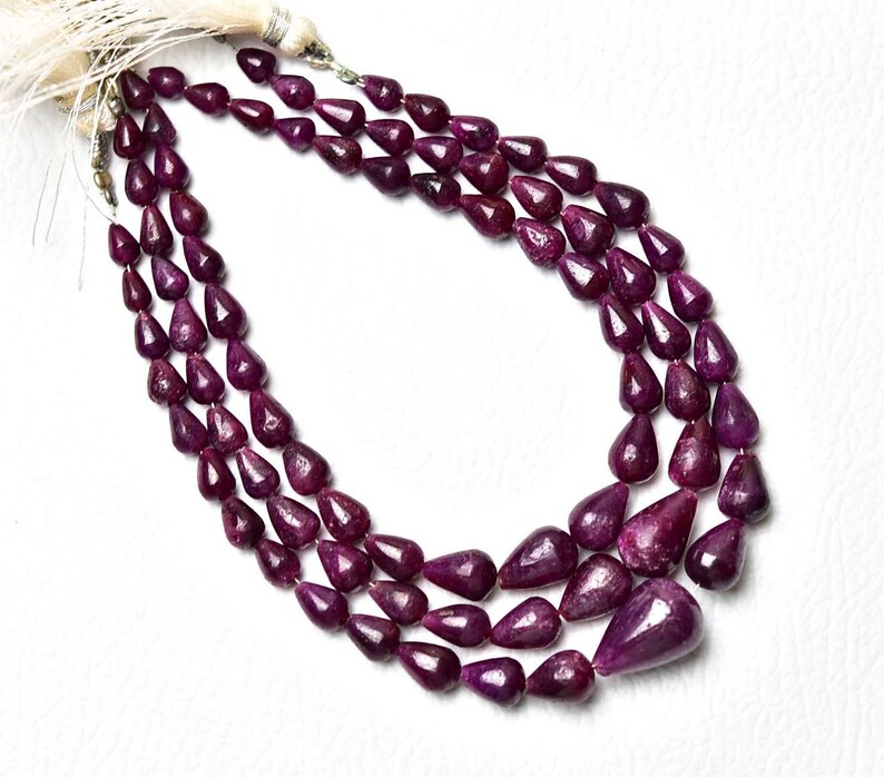 Op de afbeelding: Een ketting met drie strengen bestaande uit diep magenta, peervormige edelsteenkralen. De kralen zijn gefacetteerd en in een trapsgewijs patroon gerangschikt, bovenaan vastgezet met een lichtgekleurd kwastje en metalen onderdelen voor een gelaagd effect.