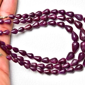 Op de afbeelding: Een ketting met drie strengen bestaande uit diep magenta, druppelvormige kralen met een gladde, gepolijste afwerking. De kralen zijn geregen aan een dunne draad en aan de uiteinden vastgezet met witte kwastjes voor een gelaagd effect.