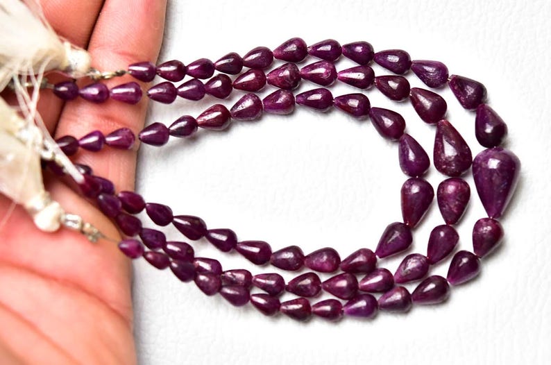 Op de afbeelding: Een ketting met drie strengen bestaande uit diep magenta, druppelvormige kralen met een gladde, gepolijste afwerking. De kralen zijn geregen aan een dunne draad en aan de uiteinden vastgezet met witte kwastjes voor een gelaagd effect.