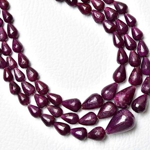 Op de afbeelding: Een ketting met drie strengen bestaande uit diep magenta, traanvormige kralen met een gepolijste afwerking. De kralen zijn in een trapsgewijs patroon gerangschikt, met de grootste traanvormige kraal in het midden onderaan de binnenste streng.