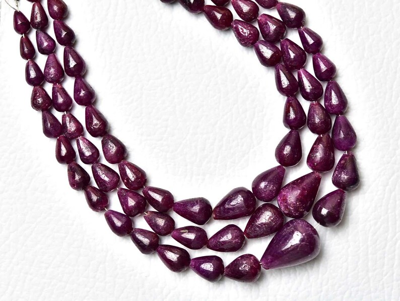 Op de afbeelding: Een ketting met drie strengen bestaande uit diep magenta, traanvormige kralen met een gepolijste afwerking. De kralen zijn in een trapsgewijs patroon gerangschikt, met de grootste traanvormige kraal in het midden onderaan de binnenste streng.