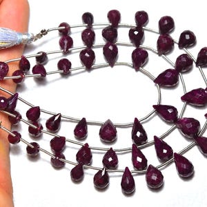 Puede incluir: Primer plano de hilos de piedras preciosas de rubí rojo oscuro en varias formas, incluyendo lágrimas y cuentas facetadas. Las piedras están ensartadas en alambre de plata, con borlas azul claro. Ideal para la fabricación de joyas.