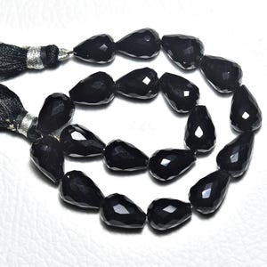 Peut inclure: Un fil de perles en onyx noir en forme de goutte, facettées. Les perles sont enfilées, avec un pompon noir à chaque extrémité. Les perles ont une finition brillante et sont disposées en cercle.