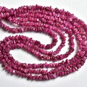 Op de afbeelding: Een lange streng van kleine, onregelmatige, ruwe robijn edelsteensplinters in een levendige magenta-roze kleur. De stenen zijn geregen aan een dunne draad, wat zorgt voor een getextureerd, gelaagd uiterlijk met een natuurlijke, ongepolijste afwerking.