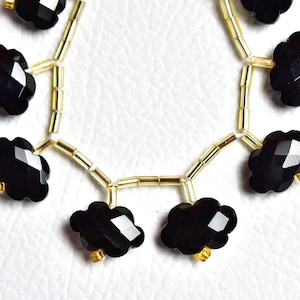 Peut inclure: Un collier de perles noires à facettes en forme de nuages, alternées avec de petites perles intercalaires cylindriques de couleur or métallique. Les perles noires présentent une surface à facettes qui reflète la lumière, disposées en ligne courbe.