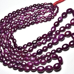 May include: A strand of deep red ruby beads, oval shaped, with a smooth, polished finish. The beads are strung on a thin cord and are arranged in a loose, flowing pattern.
