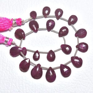 Peut inclure: Gros plan sur des rangées de perles de pierres précieuses en rubis rouge foncé facettées en forme de goutte d'eau. Les perles sont enfilées sur un fil d'argent. Deux pompons roses vifs sont visibles. Idéal pour la création de bijoux.