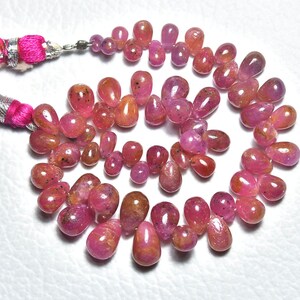 May include: A strand of pink ruby gemstone beads in a teardrop shape. The beads are strung on a cord and have a pink tassel on each end.