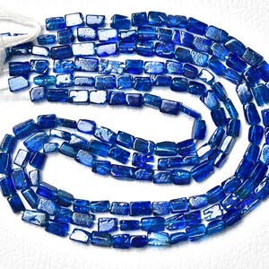 May include: A strand of rectangular, deep blue kyanite beads arranged in a coiled pattern. The beads feature a polished, reflective surface with visible internal textures and variations in color intensity, strung together on a white thread.
