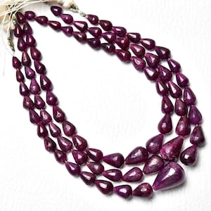 Op de afbeelding: Een ketting met drie strengen van diep magenta, peervormige robijnkralen. De kralen zijn gefacetteerd en gerangschikt in een trapsgewijs druppelpatroon, vastgezet aan de bovenkant met wit koord. De stenen hebben een rijke, verzadigde kleur.