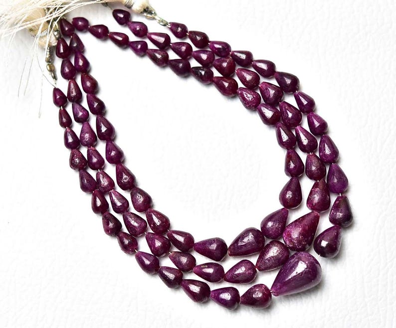 Op de afbeelding: Een ketting met drie strengen van diep magenta, peervormige robijnkralen. De kralen zijn gefacetteerd en gerangschikt in een trapsgewijs druppelpatroon, vastgezet aan de bovenkant met wit koord. De stenen hebben een rijke, verzadigde kleur.