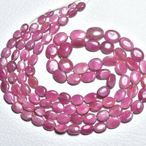 Könnte beinhalten: Stränge aus oval geformten, polierten Rubin-Edelsteinen in verschiedenen Rosa- und Rottönen. Die Edelsteine sind aufgefädelt und bilden eine Halskette oder einen Strang. Die Perlen haben eine glatte, glänzende Oberfläche und sind in einem fließenden Muster angeordnet.