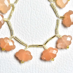 May include: A strand of faceted, peach-coloured, cloud-shaped beads interspersed with small, metallic gold-toned cylindrical spacer beads. The beads are arranged in a curved formation, showcasing a shimmering, translucent quality and a polished finish.