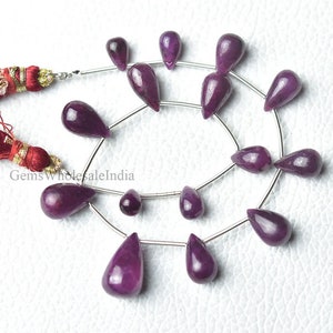 May include: A collection of polished, teardrop-shaped, deep purple gemstones strung on silver wire. Two burgundy tassels with gold accents are visible in the upper left corner. The gemstones vary in size and are arranged in a circular pattern.