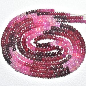 Pode incluir: Um fio de contas de pedras preciosas facetadas em tons de rosa e vermelho, dispostas em espiral. As contas variam em cor, do bordô escuro ao rosa claro, criando um efeito degradê. As contas são enfiadas em um fio branco.
