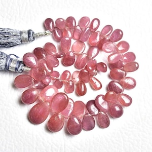 May include: A strand of polished, pear-shaped pink tourmaline gemstones arranged in a circular pattern. The translucent, faceted stones exhibit varying shades of deep rose and magenta, attached to a pair of grey silk tassels with silver thread.