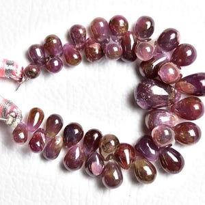May include: A strand of teardrop-shaped, polished gemstones in shades of deep magenta, burgundy, and dark purple, arranged in a circular pattern and attached to two light pink silk tassels with silver-colored metallic thread bindings.