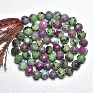 May include: A strand of green, pink, and black gemstone beads. The beads are faceted and have a speckled appearance. The strand is finished with a red and green tassel.