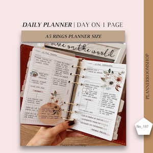 May include: A close-up of an open A5 planner with gold rings. The planner is open to a daily spread with the date "22 Aug Tuesday" and "23 Aug Wednesday" written at the top of each page. The left page has a list of tasks and the right page has a list of tasks and a floral sticker. The planner is labeled "Daily Planner | Day on 1 Page" and "A5 Rings Planner Size".