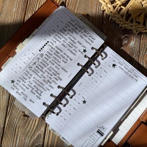 May include: A brown leather ring-bound planner open to February 2026, featuring white pages with handwritten daily entries, black ink text, star icons, and tabbed dividers. The planner displays a monthly calendar layout with space for notes.