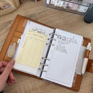 May include: A brown leather planner is open to a page with a handwritten list and a yellow index card. A white pen rests on the right side. Decorative tapes and a small bag are visible in the background.
