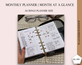 Puede incluir: Un planificador A6 abierto con anillas doradas. El planificador está abierto en el mes de julio y tiene una vista de calendario mensual. El calendario tiene notas escritas a mano y pegatinas. El planificador está sobre una superficie estampada.