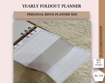 Può includere: Un'agenda annuale pieghevole, formato per planner ad anelli personali, con un layout a griglia per la pianificazione. Il testo "YEARLY FOLDOUT PLANNER" e "PERSONAL RINGS PLANNER SIZE" sono visibili. L'agenda è su una superficie strutturata.