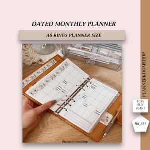 May include: A tan leather A6 ring planner featuring a monthly calendar layout with floral washi tape accents. The planner includes white pages with black grid lines, a metal ring mechanism, and a pen holder. Text includes DATED MONTHLY PLANNER, A6 RINGS PLANNER SIZE, PLANNERROOMSHOP, MON SUN START, and No_311.