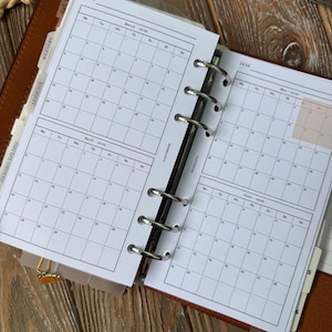 May include: Open planner with calendar pages for March, April, May, and June 2026. The pages are white with black grid lines and dates. The planner is brown with silver rings and a gold-colored clip.