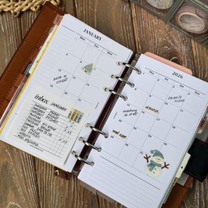 Puede incluir: Agenda abierto con páginas del calendario de enero. El calendario incluye pegatinas de un cactus y un muñeco de nieve. Una lista de tareas está en la página izquierda. La agenda es marrón con anillos plateados y un fondo de madera.