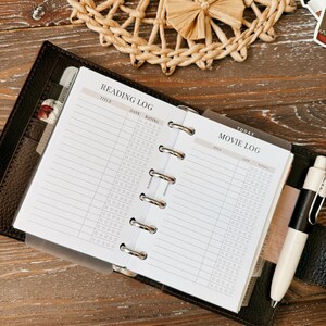 May include: A black leather ring-bound planner open to pages labeled Reading Log and Movie Log. The white pages feature columns for title, date, and star ratings. A white and black pen is tucked into the side, and a translucent divider is visible.