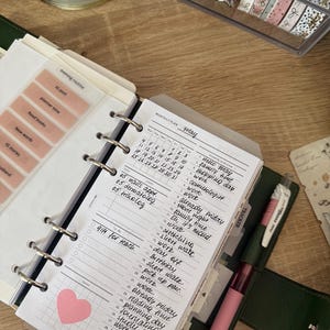 May include: A ring-bound planner open to a monthly calendar page for May with handwritten entries. The page features a grid layout, a pink heart sticker, and a section for daily tasks. A multi-colored pen rests on the dark green cover next to the planner.