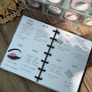 May include: A planner notebook with a weekly schedule, featuring handwritten notes and a black binding. Decorative washi tape rolls are visible in a clear storage container in the background. The notebook is open on a wooden surface.