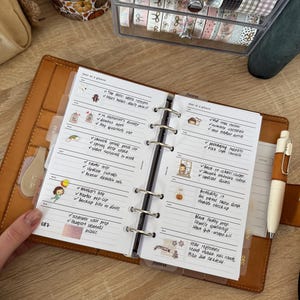 Może przedstawiać: Otwarty brązowy planer z odręcznymi notatkami i naklejkami. Planer zawiera listę zadań i spotkań. Pióro spoczywa po prawej stronie planera. Planer leży na drewnianej powierzchni.