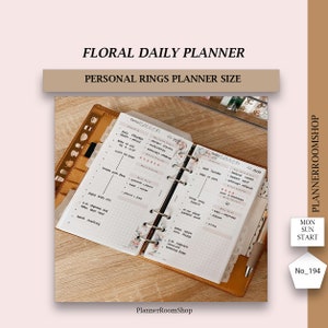 Puede incluir: Una agenda diaria floral con una cubierta de cuero marrón y anillos plateados. La agenda está abierta en una página con un diseño de cuadrícula y las palabras "Daily Planner" y "Weekly Planner" en la parte superior. La agenda está decorada con pegatinas florales.