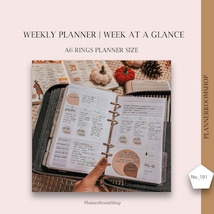 Peut inclure: Un planneur A6 ouvert sur une semaine, avec une vue d'ensemble de la semaine. Le planneur est ouvert sur la semaine du 5, avec les jours de la semaine étiquetés. Le planneur a une couverture noire et des anneaux dorés. Les pages sont remplies de notes manuscrites et d'autocollants. Le planneur est entouré de décorations sur le thème de l'automne, notamment des citrouilles et des pommes de pin.