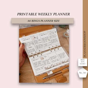 Könnte beinhalten: Ein A6-großer, druckbarer Wochenplaner mit goldenen Ringen. Der Planer ist auf eine Wochenübersicht geöffnet, mit handschriftlichen Notizen und Aufklebern. Der Planer ist aus braunem Leder mit dem aufgedruckten Text "PlannerRoomShop" auf der Vorderseite.