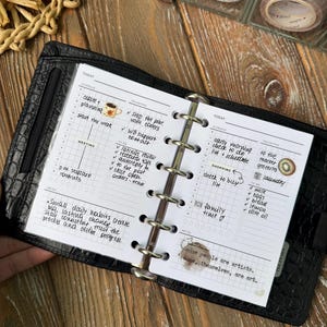 Könnte beinhalten: Ein schwarzer Lederplaner, der auf einer Seite mit Tagesplan und To-Do-Listen geöffnet ist. Handschriftliche Notizen und eine Kaffeetassen-Illustration sind zu sehen. Dekorative Klebebandrollen sind im Hintergrund.
