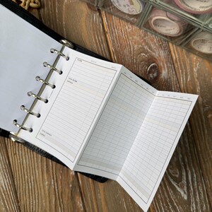 May include: A black binder-style planner with open pages. The planner has a white insert with a financial planning layout, including sections for income and expenses. Decorative washi tape rolls are visible in the background.